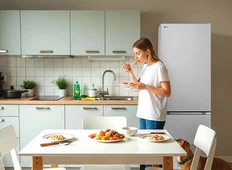 White LG small-capacity fridge freezer in a modern kitchen. A woman eats breakfast at a table with food, while a dog stands behind her looking up.