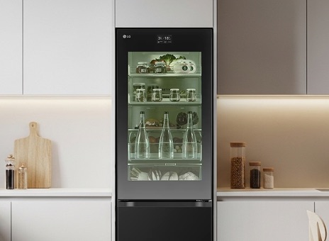 LG tall fridge with a see through glass door in a minimalist kitchen. The interior is lit with green lighting, clearly showing stored fresh groceries and beverages.