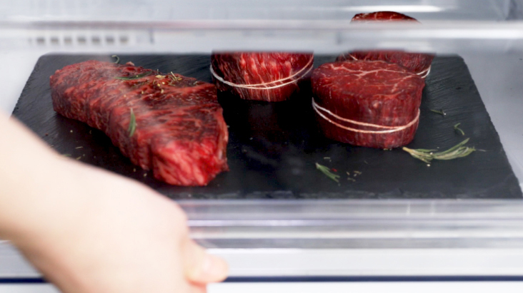Hand placing a tray of seasoned raw beef steaks, tied with string, into a refrigerator.