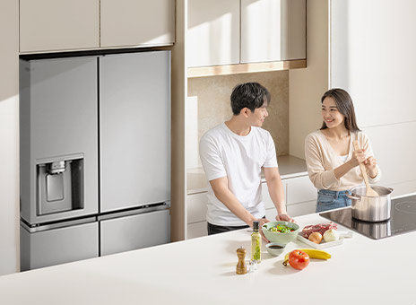 A large stainless LG refrigerator freezer with spacious storage in a stylish kitchen. A man and a woman cook in front of a table.	