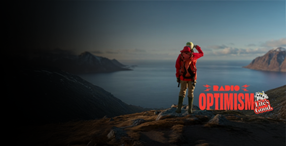 A woman wearing a red hiking jacket is standing in a place where the vast ocean and snow-covered mountains can be seen. The image also features the "Radio Optimism" slogan logo and the "Life's Good." logo alongside the woman.	