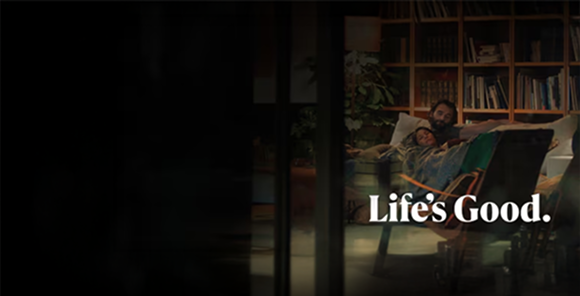 A man is sitting on a sofa in front of a bookshelf. A woman is leaning against the man's arm. The text Life's Good is written beneath the image.