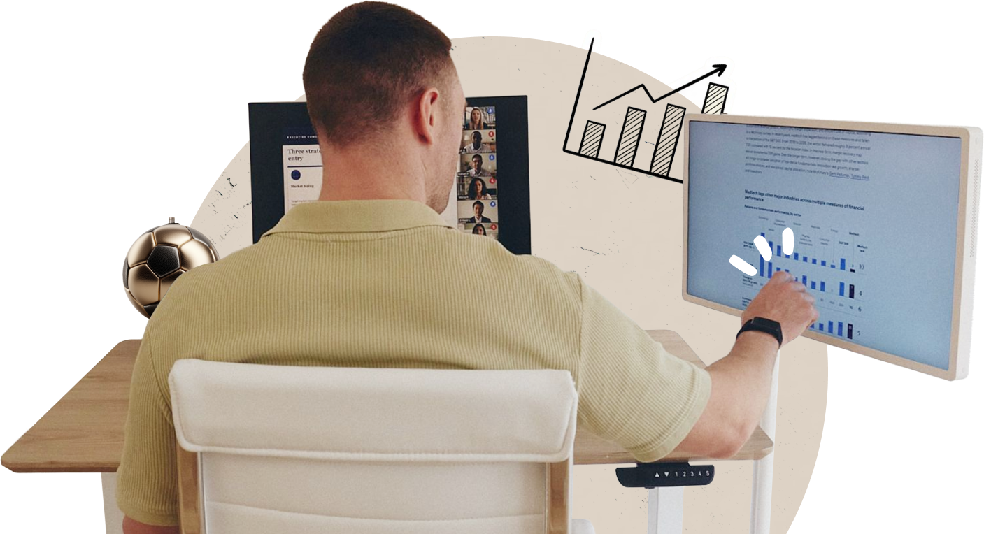 The back of a man touching a StanbyME 2 smart screen displaying data, next to a laptop showing a video meeting.