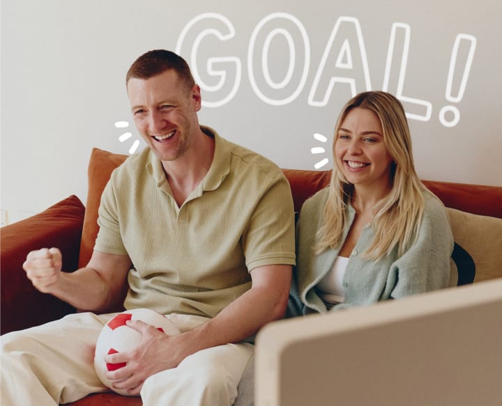 An excited man sitting on a sofa and holding a soft soocer ball toy in one hand, with a woman laughing alongside him, with 'GOAL!' text.
