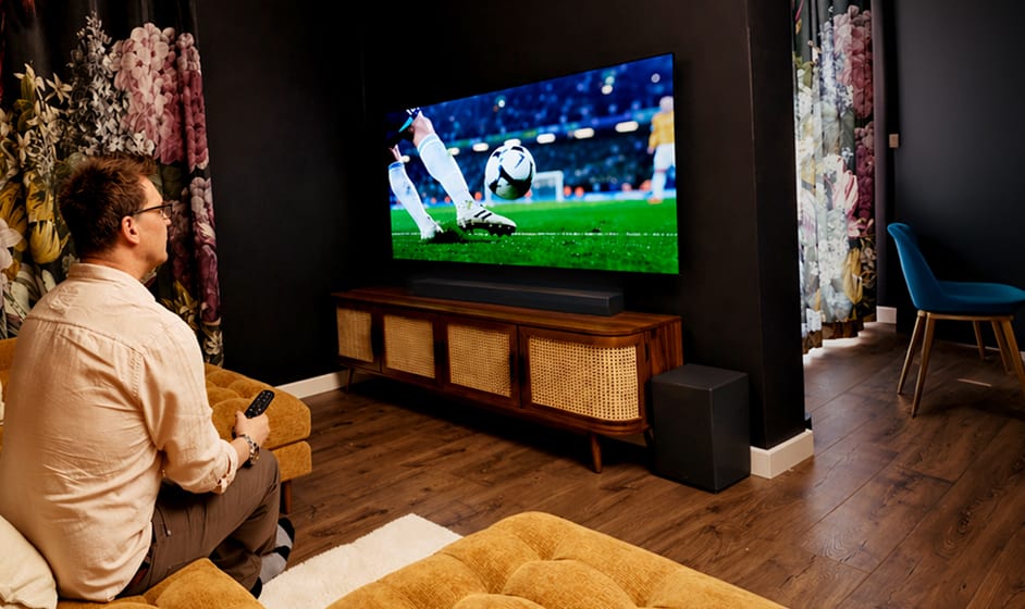 A man sits on a sofa in a bright living room, holding a game controller while watching a soccer match on a wall-mounted TV.