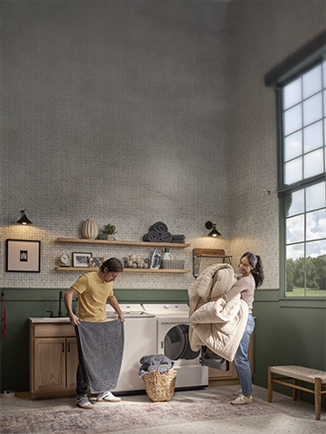 A cheerful couple does laundry together using their LG top load washer and dryer in a bright, stylish laundry room. They’re smiling while folding towels and unloading a comforter, showing how easy and enjoyable laundry can be with reliable LG appliances—ideal for illustrating LG service and support.