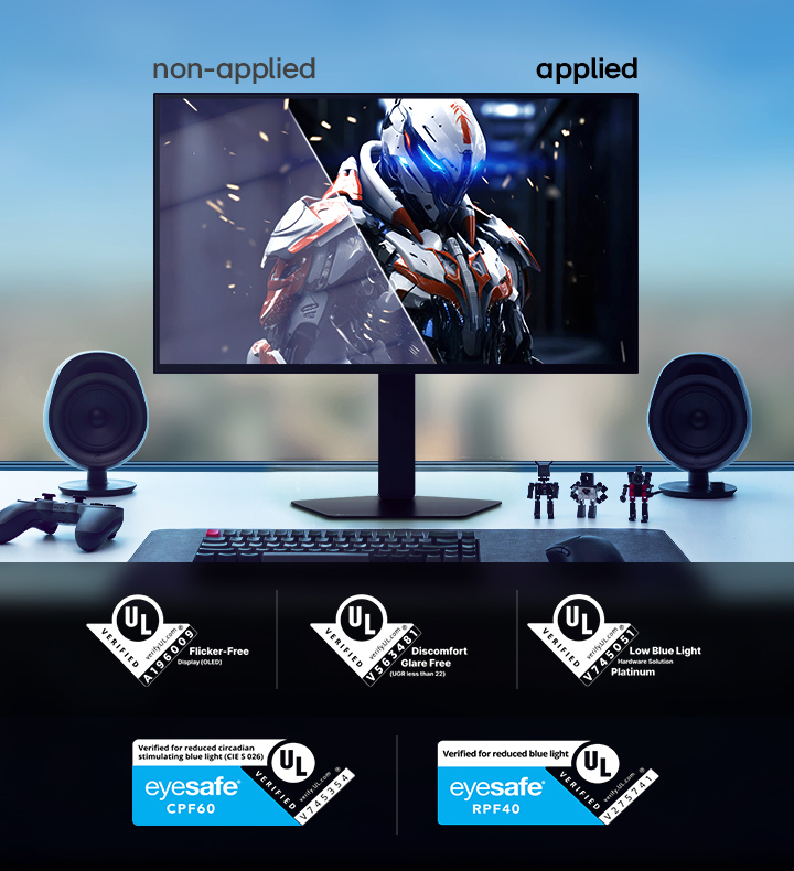 LG UltraGear 27GX790B monitor on a desk setup displaying a side-by-side comparison labeled ‘non-applied’ and ‘applied,’ showing different visual states of a sci-fi armored character. The desk includes speakers, a keyboard, figurines, a controller, a PC tower, and headphones. Along the bottom are multiple UL certification badges for flicker-free, discomfort glare-free, low blue light, and Eyesafe credentials