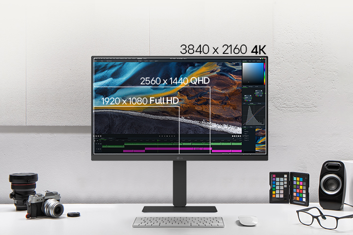 Front view of a white LG monitor on a desk, displaying layered screen resolutions for comparison, with cameras, books, color chips, and speakers arranged around it.