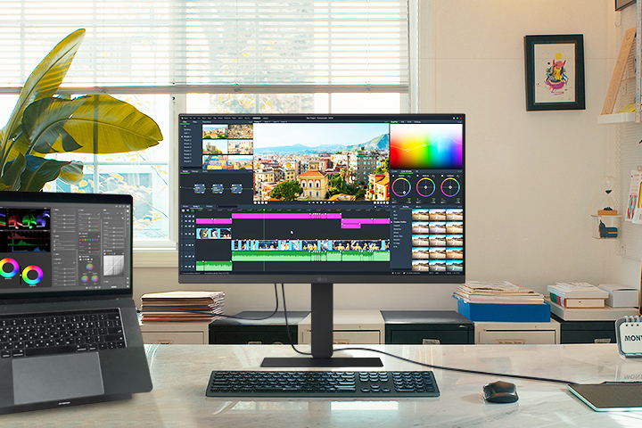 A creative workspace featuring an LG monitor and a connected laptop, both displaying a video editing interface with color grading tools and a timeline.