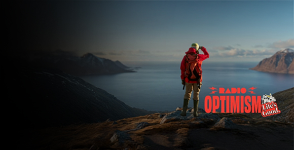 A woman wearing a red hiking jacket is standing in a place where the vast ocean and snow-covered mountains can be seen. The image also features the "Radio Optimism" slogan logo and the "Life's Good." logo alongside the woman.