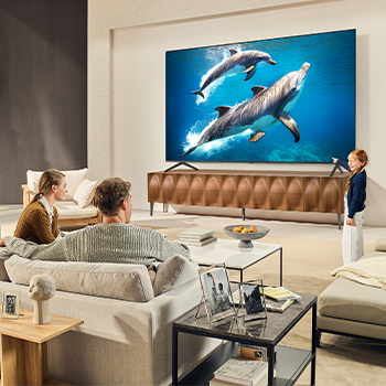Une famille est assise sur un canapé face à un LG QNED TV placé au-dessus d’un meuble TV. Une petite fille pointe du doigt un écran représentant deux dauphins. Le titre explique comment profiter de la haute résolution sur un écran géant.