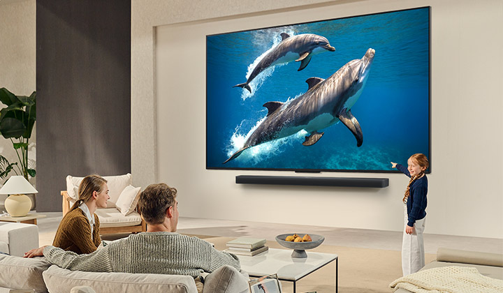 Une famille est assise sur un canapé face à un LG QNED TV monté sur un mur. Une petite fille pointe du doigt un écran représentant deux dauphins. 