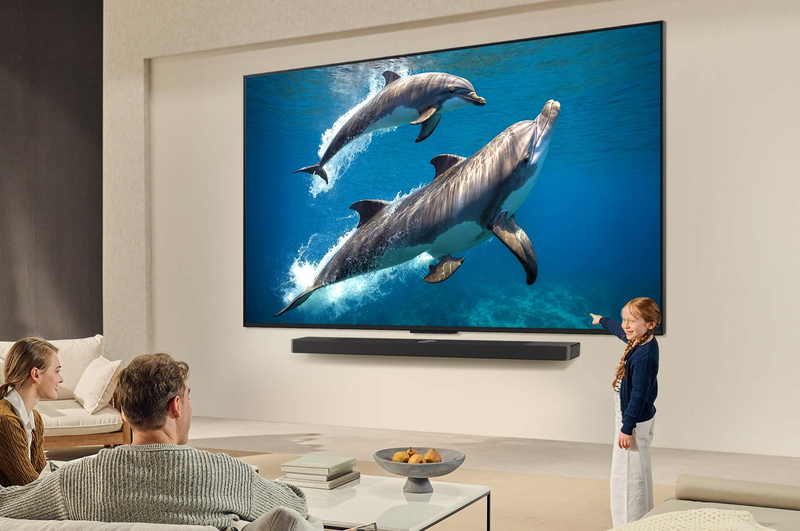Une famille est assise sur un canapé face à un LG QNED TV monté sur un mur au-dessus d’un LG Soundbar. Une petite fille pointe du doigt un écran représentant deux dauphins. Le titre met en avant comment chaque action devient encore plus captivante sur un écran géant.