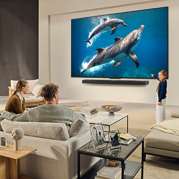 Une famille est assise sur un canapé face à un LG QNED TV monté sur un mur au-dessus d’un LG Soundbar. Une petite fille pointe du doigt un écran représentant deux dauphins. Le titre met en avant comment chaque action devient encore plus captivante sur un écran géant.