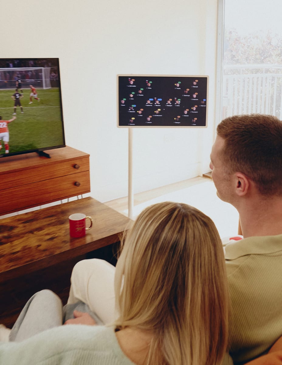 Le dos d’un homme et d’une femme assis côte à côte sur un canapé, regardant un match de football à la TV tout en consultant une analyse du match sur un écran intelligent StanbyME 2 .