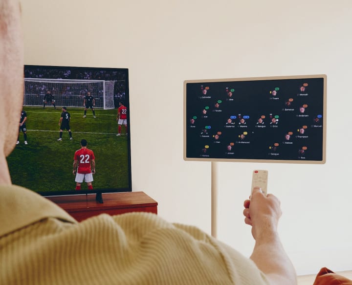 Le dos d’un homme pointant une télécommande vers une TV diffusant un match de football et un écran intelligent StanbyME 2 affichant une analyse du match.