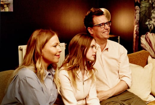 Une famille de trois personnes est assise tout près sur un sofa, regardant quelque chose hors champ avec des expressions attentives, comme si elle regardait la TV.