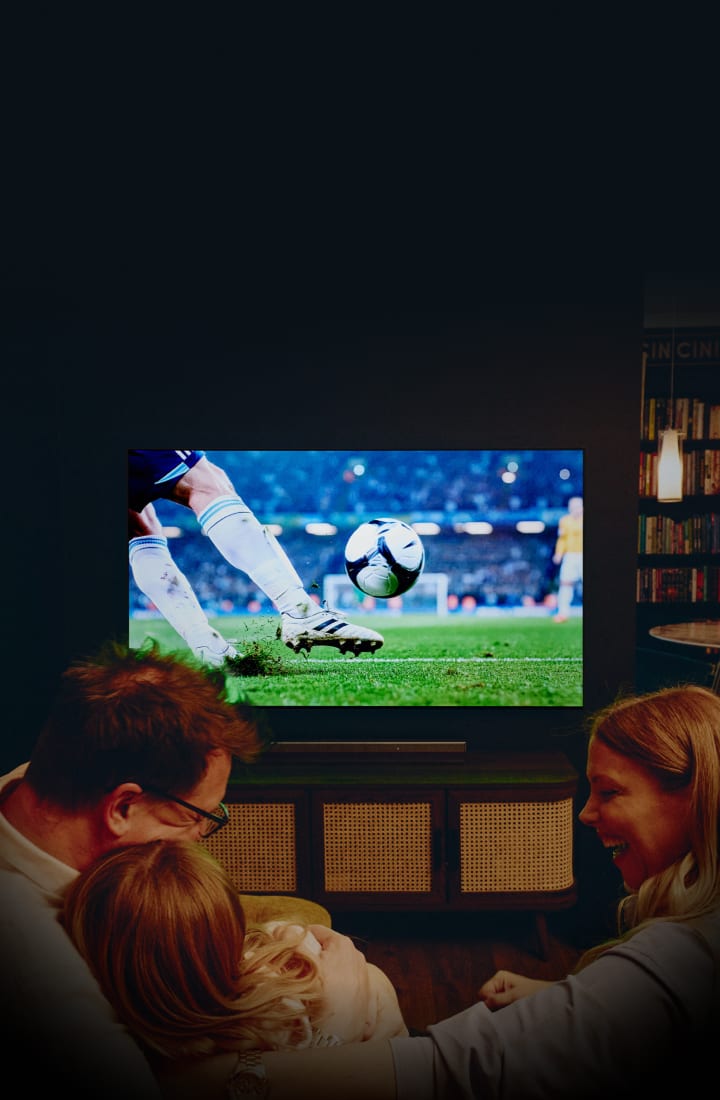 Une famille dans un salon confortable regarde un match de soccer sur une grande TV murale montrant un joueur frappant un ballon dans un stade, avec une lumière chaleureuse et une ambiance cinématographique.