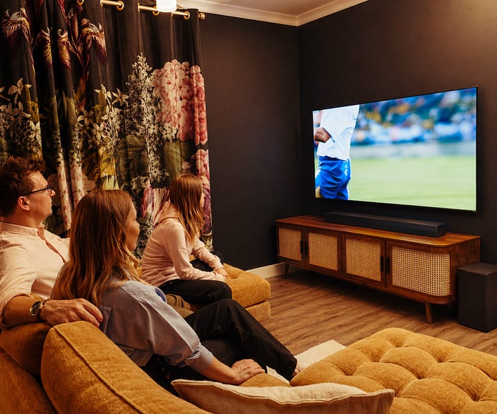 Une famille de trois personnes est assise ensemble sur un sofa dans un salon confortable et faiblement éclairé, regardant un match de soccer sur une grande TV LG OLED. La pièce présente des murs foncés, des rideaux à motif floral, une console média en bois et des haut-parleurs, créant une atmosphère chaleureuse et intime.