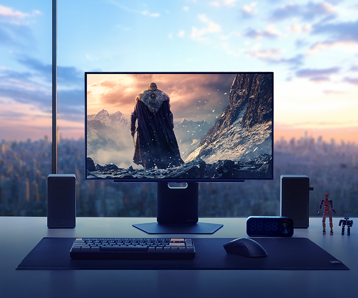 Un moniteur de jeu haut de gamme affiche une scène de jeu fantastique spectaculaire mettant en scène un guerrier encapuchonné devant une chaîne de montagnes enneigées. Le moniteur est posé sur un bureau élégant dans un appartement en hauteur, avec une vue sur la skyline de la ville au coucher du soleil en arrière-plan. Autour du moniteur se trouvent des enceintes, un clavier, un casque, une horloge et de petites figurines, illustrant une configuration de jeu immersive et stylée.