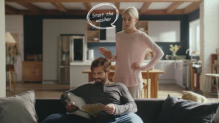 Dans une cuisine moderne et intelligente, une femme utilise une commande vocale pour démarrer la machine à laver avec LG ThinQ IA, tandis qu'un homme lit sur le canapé en arrière-plan.