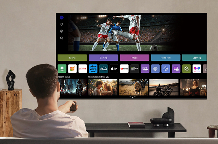 شخص على الأريكة يحمل جهاز تحكم عن بُعد. تلفزيون LG AI TV على الحائط مع LG webOS على الشاشة.