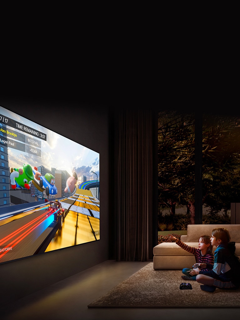 منظر جانبي لـ LG TV كبير جدًا معلق على الحائط في غرفة معيشة مريحة يعرض لعبة ملونة، وطفلان يجلسان على الأرض أمام التلفزيون ويحملان أجهزة التحكم في الألعاب.