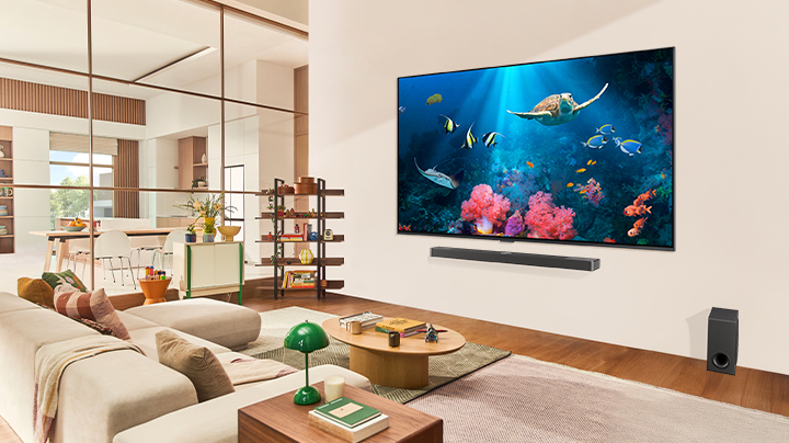 عائلة في غرفة معيشة مع LG TV كبير للغاية معلق على الحائط، يعرض مشهدًا للمحيط يتضمن مرجانًا وسلحفاة على الشاشة.