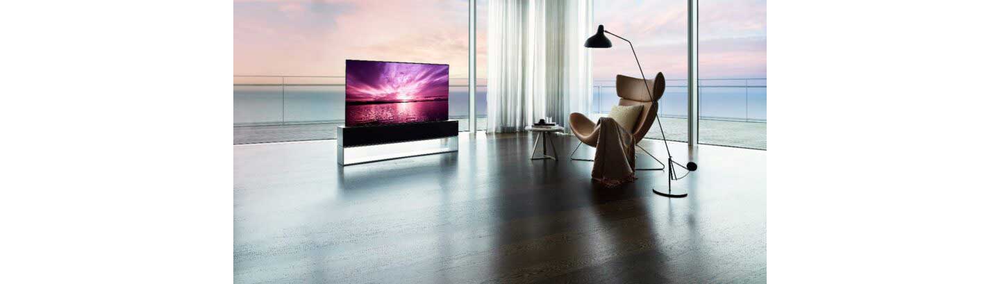 LG SIGNATURE OLED R inside a bright modern space with expansive windows in Full View Mode, a its true-to-life color displays a purple sunset that harmonizes with the coastal view outside