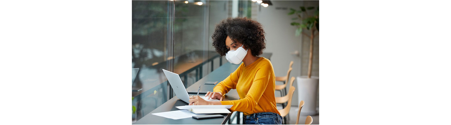 A woman wearing LG PuriCare™ Wearable Air Purifier as she works on a laptop at a cafe to protect herself during uncertain times.