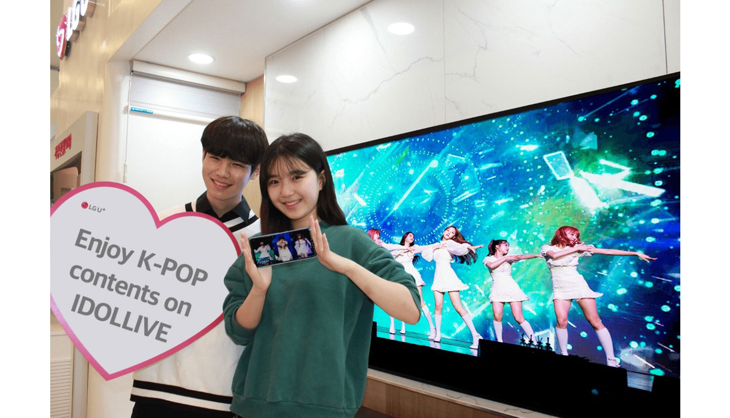 A man and woman posing in front of an LG Smart TV displaying a K-pop group’s performance to celebrate more K-culture content coming to LG’s webOS platform.