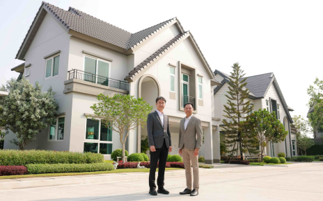 Two men stand on the street in front of a large home with surrounding greenery.
