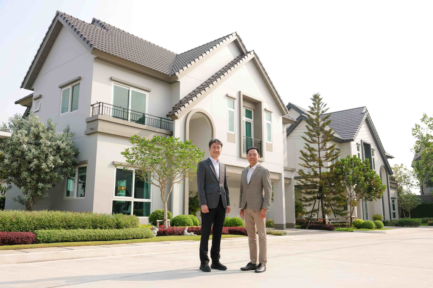 Two men stand on the street in front of a large home with surrounding greenery.
