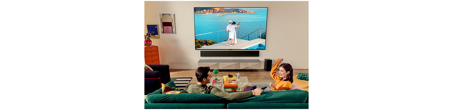 A man and a woman sitting on a green sofa and having fun while watching a TV displaying a couple dancing