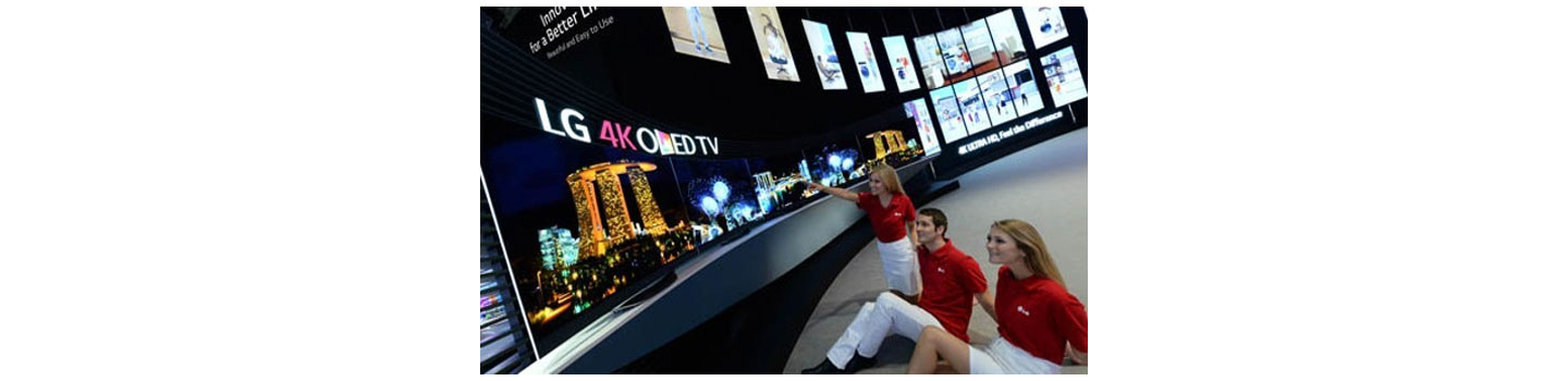 Three models sit down on the floor and look up to the world's first LG 4K curved OLED TV products that are positioned on a display stand.