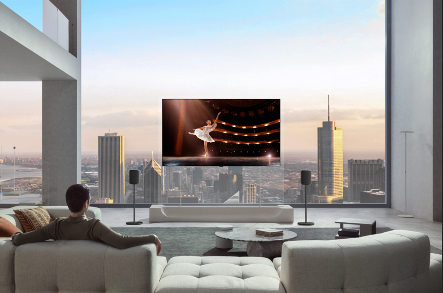  	A man is sitting on a sofa in his city apartment, watching a ballerina performance on an LG OLED TV mounted on a transparent stand with two speakers.