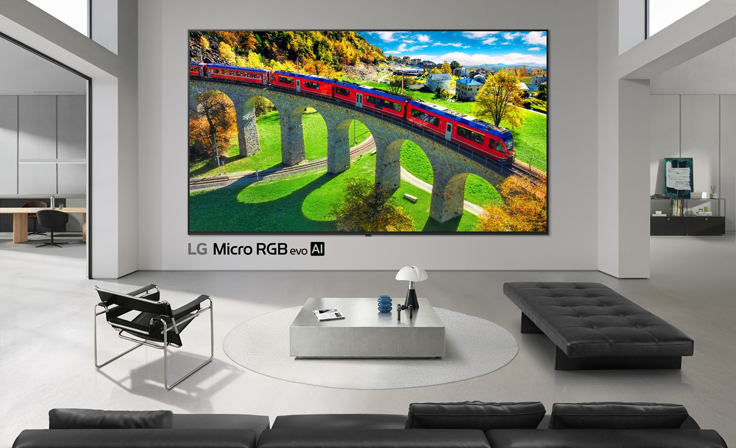 An ultra-large LG Micro RGB evo TV is mounted to the wall in a modern living room, with a vivid red train crossing a stone bridge displayed on the screen.