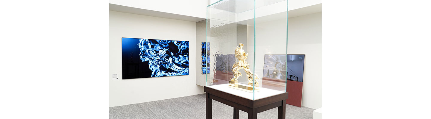 A gold sculpture inside a glass display case with an LG OLED TV and Objet Easel displaying artwork in the background