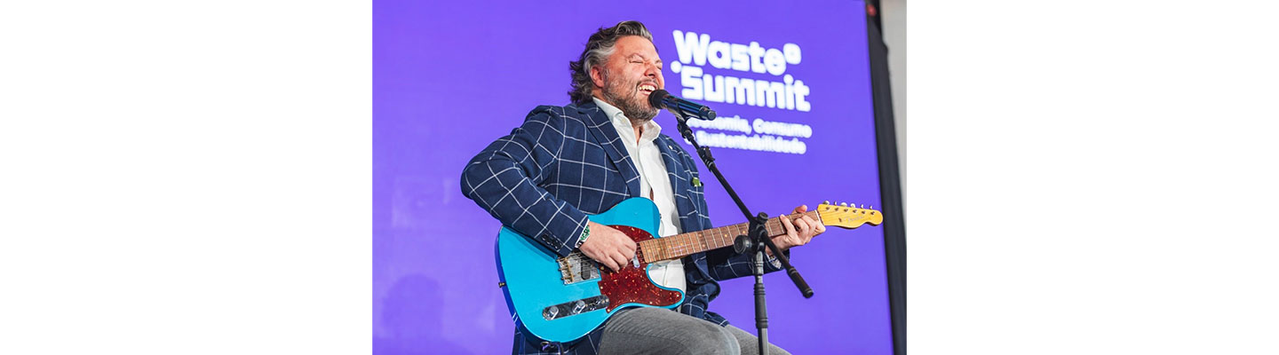 Comedian César Mourão playing a guitar and singing on stage during the Waste Summit.