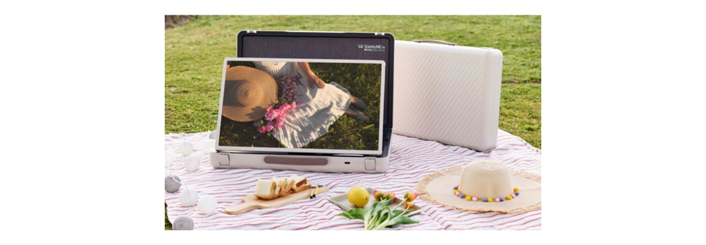 A blanket is spread out on a grassy lawn for a picnic with the LG StanbyME Go model in landscape mode displaying the image of a woman