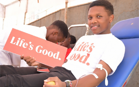 A blood donor wearing Life's Good T-shirt posing for a photo while holding a panel that also reads 