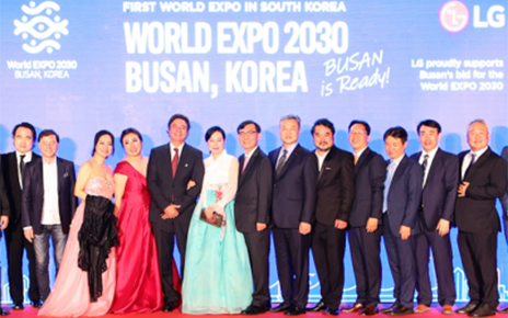 A photo of opera-related actors and staff posing on the podium with a digital screen at the back displaying the phrase 