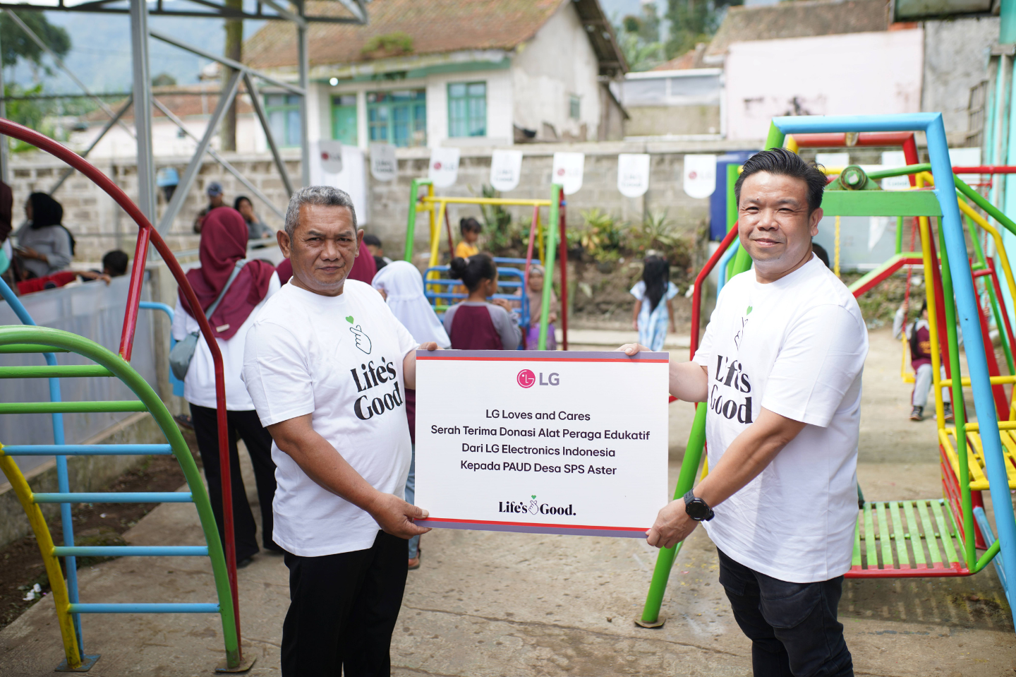Two men are holding a sign that reads "LG Loves and Cares," with children playing on a playground in the background.