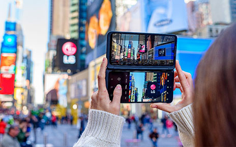 A woman holds LG G8XThinQ with LG Dual Screen in her two hands vertically to check the weather on the screen at an urban space with buildings.