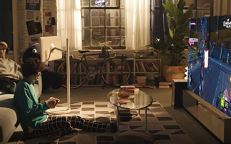 One friend plays a video game on LG’s latest OLED TV while the other lays on the living room sofa next to LG StanbyME.