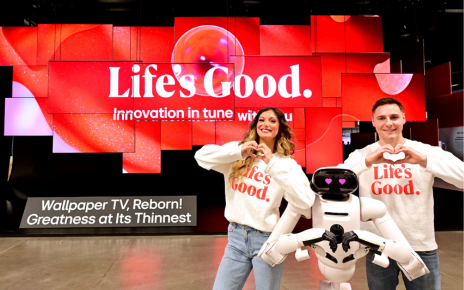 A man and woman pose with the LG CLOiD home robot, all making a heart shape with their hands in front of the In Tune Monument at the booth entrance.