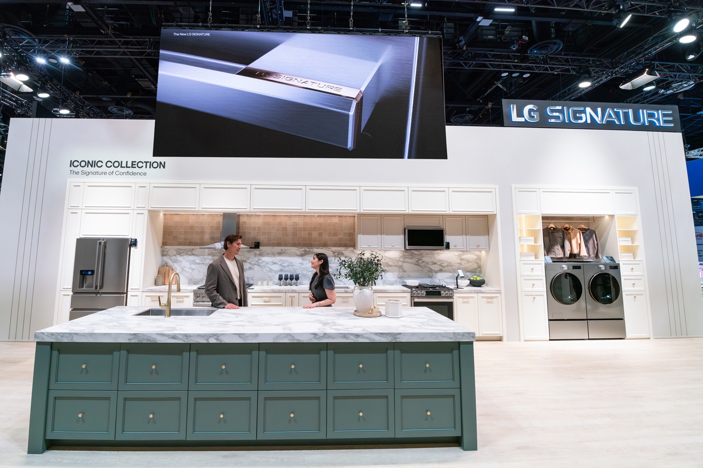 A man and a woman stand behind a green kitchen island in the LG SIGNATURE Iconic Collection display at LG’s booth during KBIS 2026, with a variety of LG SIGNATURE appliances in a kitchen setup behind them.
