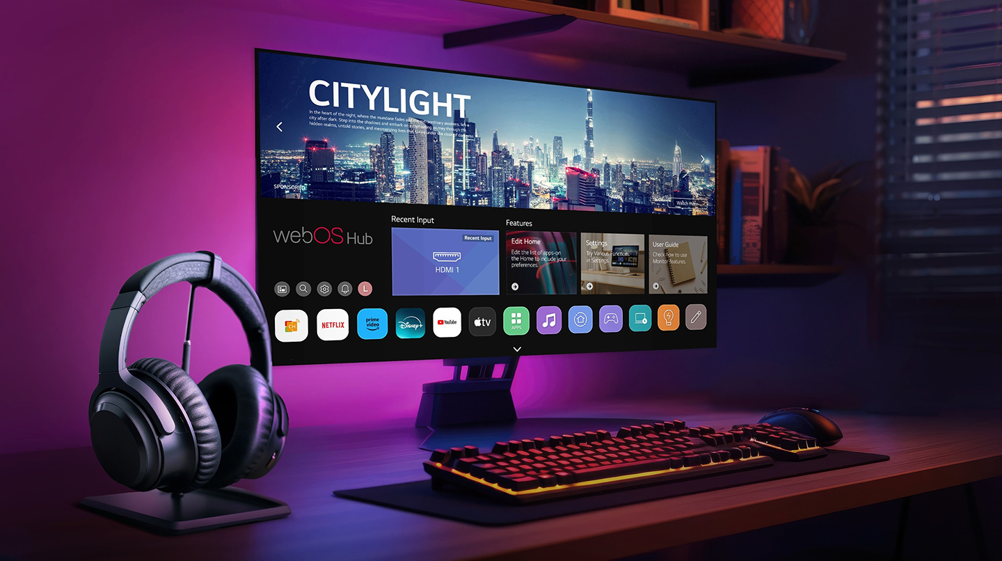 The monitor of a gaming setup displays the webOS Hub platform, with wireless headphones placed beside the keyboard