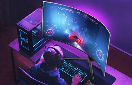 Man sits a desk with curveed monitor playing computer games. He has a brighlty lit gaming station set-up.