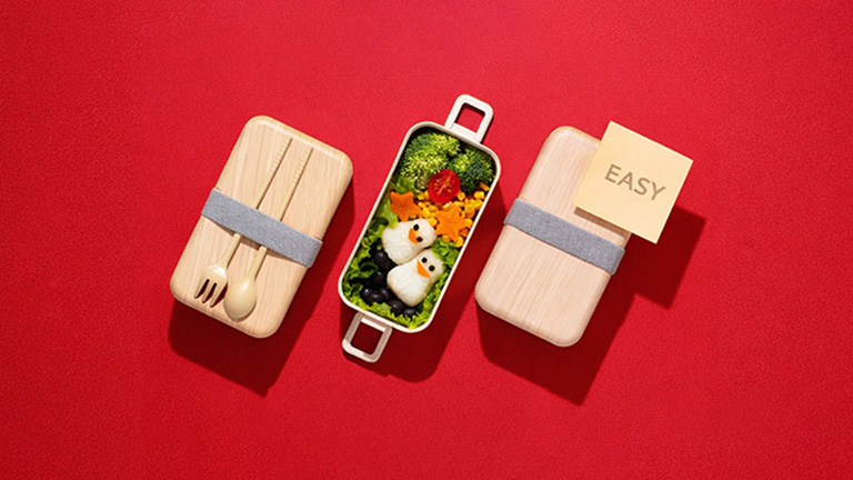 An image showing an easy bento box meal with penguin-shaped rice balls and colorful vegetables on a red background, illustrating a simple meal-prep concept.	